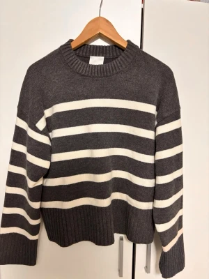 Gråvit randig stickad tröja H&M - Säljer en grå stickad tröja från H&M med breda vita horisontella ränder. Tröjan har rund halsringning, ribbade muddar och avslut. Perfekt för dig som gillar en avslappnad och trendig stil.