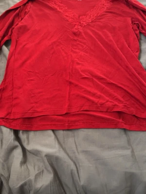 Röd långärmad topp med spets - Snygg röd långärmad topp med v-ringning och spetsdetaljer längs halsen. Tillverkad i mjukt material som känns skönt mot huden. Perfekt att styla med jeans eller kjol för en trendig look.