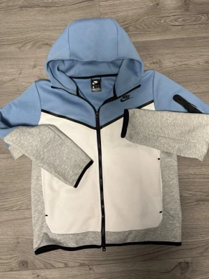 Nike hoodie i blått, vitt och grått - Snygg hoodie från Nike i en fräsch färgkombination av ljusblått, vitt och grått. Dragkedja framtill, huva och svarta detaljer längs kanterna. Nike-logga på bröstet och fickor på sidorna. Perfekt för en sportig och avslappnad stil.