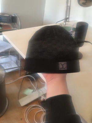 Svart mössa från Louis Vuitton - Snygg svart mössa från Louis Vuitton med diskret rutmönster och LV-logga framtill. Tillverkad i mjukt material som känns bekvämt på huvudet. Perfekt accessoar för att lyfta din outfit med en exklusiv touch.