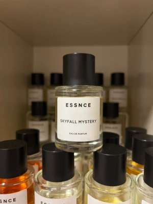 ESSNCE Skyfall Mystery parfym - Upptäck ESSNCE Skyfall Mystery Eau de Parfum i en stilren, rund glasflaska med svart lock. Parfymen har en minimalistisk vit etikett med svart text och transparent vätska. Perfekt för dig som gillar moderna och enkla designer.