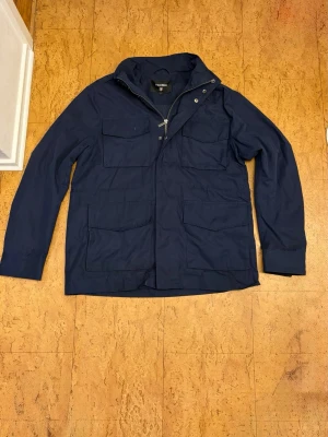 Mörkblå vindjacka från Strömberg - Stilren mörkblå vindjacka från Strömberg med fyra stora fickor framtill, hög krage och dragkedja med tryckknappar. Jackan har en rak passform och är tillverkad i ett lätt, vindtätt material. Perfekt för blåsiga dagar och enkel att matcha med olika outfits. Nypris cirka 1200kr