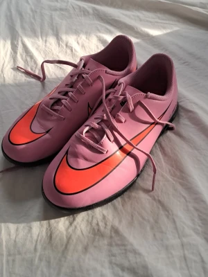 Rosa Nike fotbollsskor med orange swoosh - Säljer ett par snygga fotbollsskor från Nike i rosa med en stor orange swoosh på sidan. Skorna har snörning och låg profil, samt svart yttersula som passar för spel på konstgräs eller inomhus. Perfekta för dig som vill sticka ut på planen!