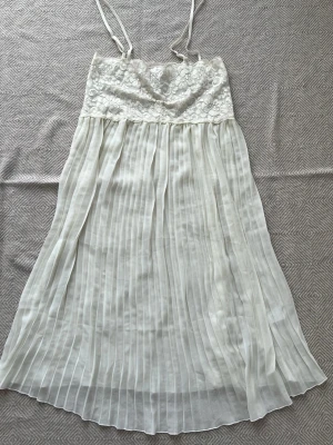 Vitt spetsnattlinne med plisserad kjol - Sött vitt nattlinne med tunna axelband, överdel i blommig spets och kjol i plisserat, skirt tyg. Perfekt för mysiga kvällar hemma. Materialet känns lätt och luftigt, och spetsen ger en romantisk touch. Nypris 799kr