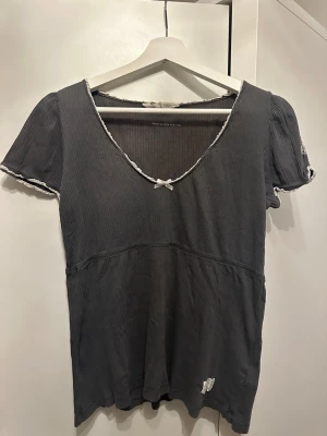 Mörkgrå topp med rosett och spets från Odd Molly - Supersöt mörkgrå topp med ribbad struktur, liten vit rosett vid urringningen och vita spetsdetaljer längs kanterna. Djup v-ringning och korta puffärmar ger en feminin vibe. Perfekt att styla med jeans eller kjol för en trendig look.