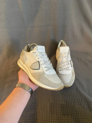 Philippe Model  - Snygga vita sneakers från Philippe Model med detaljer i beige mocka och mesh. Skorna har vita snören med logotyp, en ikonisk sköld på sidan och en bekväm sula. Perfekta för dig som gillar stilrena och trendiga sneakers.