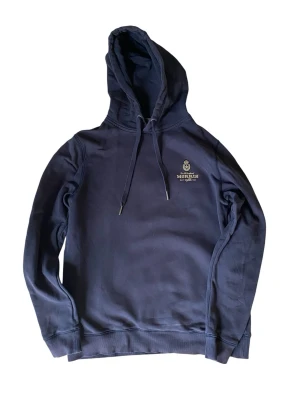 Mörkblå hoodie från Morris - Snygg mörkblå hoodie från Morris med justerbar huva och dragsnören. Diskret Morris-logga broderad på bröstet i vitt och guld. Tillverkad i mjukt bomullsmaterial och har ribbade muddar vid ärmslut och nederkant. Perfekt för en avslappnad och stilren look.