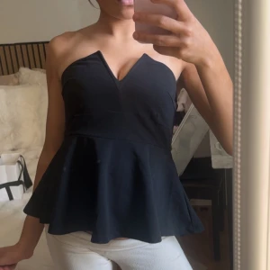 Svart axelbandslös peplum topp - Snygg svart axelbandslös peplum topp med v-ringad skärning framtill. Toppen har en figurnära passform upptill och ett utsvängt avslut nedtill som ger en feminin siluett. Perfekt att styla med jeans eller kjol för en trendig look.