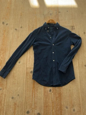 Mörkblå skjorta från Ralph Lauren - Klassisk mörkblå skjorta från Ralph Lauren i mjuk bomull. Skjortan har lång ärm, button-down krage och vita knappar framtill. Diskret broderad logga på bröstet ger en stilren touch. Perfekt till jeans eller chinos.