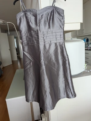 Silvergrå axelbandsklänning från Vila - Säljer en snygg grå kort klänning från Vila med tunna axelband och markerad midja. Dragkedja i sidan och resår i ryggen. Klänningen har en glansig yta och enkel, stilren design som passar perfekt till festen. 