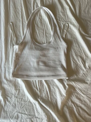 Vit ribbad croppad linnetopp - Säljer en vit ribbad croppad linnetopp med breda axelband och rund halsringning. Toppen är ärmlös och har en enkel, stilren design som passar perfekt till sommaren. Materialet är mjukt och stretchigt för en bekväm passform.