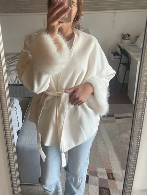 Vit kappa med fuskpälsarmar - Kort vit kappa med knytband i midjan och fluffiga fuskpälsdetaljer vid ärmsluten. Perfekt för dig som vill ha en chic och trendig look. Materialet känns mjukt och ger en lyxig känsla.