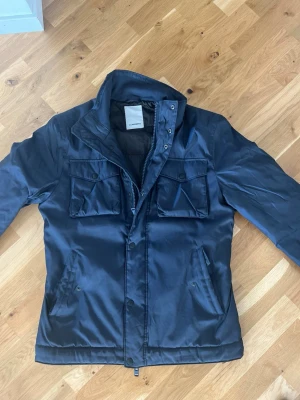 Mörkblå vindjacka från J.Lindeberg - Snygg mörkblå overshirt/varmarejacka från J.Lindeberg med hög krage, dragkedja och tryckknappar. Jackan har två bröstfickor med lock och knappar samt två sidofickor. Materialet är glansigt och vattenavvisande, perfekt för blåsiga dagar.