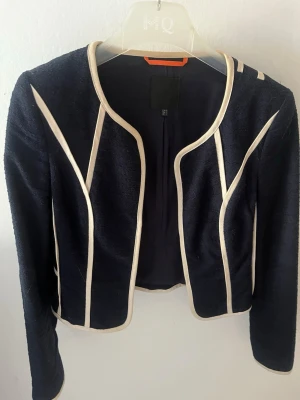 Marinblå kavaj med vita detaljer - Snygg marinblå kavaj med vita kontrastband längs sömmarna och runt kanterna. Kortare modell med rundad halsringning och långa ärmar. Perfekt för en trendig och stilren look. Materialet ser ut att vara mjukt och lätt, vilket ger en bekväm känsla.