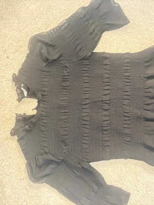 Svart smockad blus med volang - Trendig svart blus med smockad kropp och volangdetaljer vid hals och ärmslut. Tillverkad i ett tunt, transparent tyg som ger en luftig känsla. Långa puffiga ärmar med resår och volangavslut. Perfekt för dig som gillar en romantisk och chic stil.