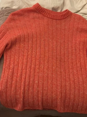 Ribbstickad orange tröja - Säljer en ribbstickad tröja i en snygg orange färg. Tröjan har rund halsringning och långa ärmar. Materialet ser mjukt och lite fluffigt ut, perfekt för kyligare dagar. Inga synliga loggor eller märken.