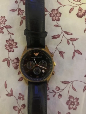 Emporio Armani svart klocka - Stilren klocka från Emporio Armani med svart urtavla och guldfärgat boett. Klockan har svart läderarmband med krokodilpräglad yta och flera små urtavlor för extra funktioner. Perfekt accessoar för dig som gillar klassisk och elegant stil.