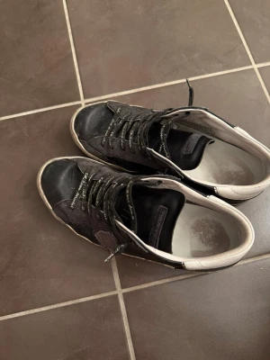 Philippe Model svarta sneakers - Snygga svarta sneakers från Philippe Model med vita detaljer på hälen och grå sula. Skorna har klassisk låg profil, svarta skosnören med text och är tillverkade i mocka och läder. Perfekta för dig som gillar stilrena och trendiga skor.