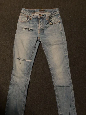 Ljusblå slitna jeans från Nudie - Säljer ett par ljusblå jeans från Nudie med slim fit och slitningar på låret och knät. Klassisk femficksmodell med orange sömmar och Nudie-logga på bakfickan. Tillverkade i mjuk denim med snygg tvätt och detaljer.