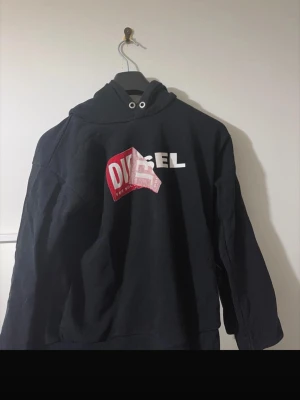 Svart hoodie från Diesel med tryck - Cool svart hoodie från Diesel med stor logga i vitt och rött framtill. Klassisk huva med dragsko och långa ärmar. Perfekt för dig som gillar streetwear och vill ha en statement-piece i garderoben.