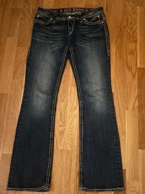 Rock Revival bootcut jeans blå - Snygga blå bootcut jeans från Rock Revival med kontrastsömmar och slitningar framtill. Jeansen har dekorativa fickor med detaljer och silverknappar. Klassisk femficksmodell i denim med cool tvätt och markerade sömmar. Säljer dom för dom var för stora på mig, storlek 29. Passar som S