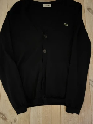 Svart cardigan från Lacoste - Klassisk svart cardigan från Lacoste med V-ringning och knäppning framtill. Tröjan har långa ärmar, ribbade muddar och den ikoniska gröna krokodilloggan på bröstet. Perfekt för en stilren och enkel look.