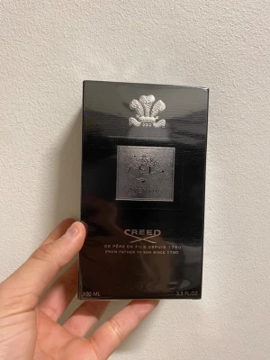 Creed Aventus Eau de Parfum 100ml - Creed Aventus Eau de Parfum i elegant svart förpackning med silverdetaljer. Flaskan rymmer 100 ml och har en lyxig design med Creed-loggan och en klassisk emblem på framsidan. En ikonisk doft som kombinerar fruktiga och träiga noter för en unik signatur.