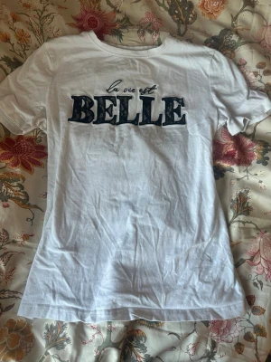 Vit t-shirt med spetsdetalj - Snygg vit t-shirt med texten 'la vie est BELLE' framtill där bokstäverna är dekorerade med svart spets. Jag vet inte vart den kommer ifrån men den är köpt i Rumänien.