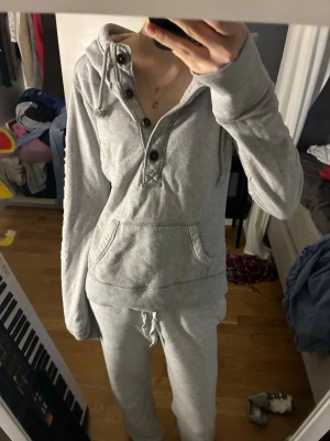 Grå hoodie från Abercrombie & Fitch - Mysig grå hoodie med knappar och huva från Abercrombie & Fitch. Stor känguruficka framtill och tryck med texten 'California' längs ena ärmen. Perfekt för chill dagar och har en avslappnad passform.