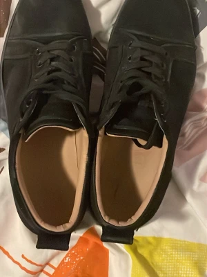 Svarta sneakers från Christian Louboutin - Snygga svarta sneakers från Christian Louboutin med den ikoniska röda sulan och diskret logga undertill. Skorna har en klassisk låg siluett och är tillverkade i svart material med matchande svarta snören. Perfekta för dig som vill sticka ut med stil.