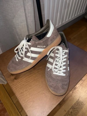 Adidas Spezial bruna sneakers - Säljer ett par Adidas Spezial sneakers i brunt mocka med vita detaljer. Skorna har vit sula och vita skosnören, samt Adidas-logga och texten 'Spezial' i vitt på sidan. Köpta på Vinted, säljer då de är för stora för mig. Har endast testat dem, och förra ägaren hade använt dem fåtal gånger. Mockan är lite tilltufsad på ena hälen, men inget som är fult eller stör!🤎