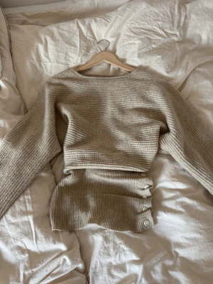 Beige ribbstickad tröja - Mysig beige ribbstickad tröja med lång ärm och rund halsringning. Tröjan har en croppad passform och är gjord i ett mjukt, stretchigt material som troligtvis är bomull eller bomullsblandning. Perfekt för lager på lager och enkel att styla till olika outfits.