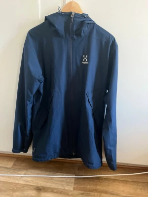 Mörkblå regnjacka från Haglöfs - Snygg mörkblå regnjacka från Haglöfs med huva och hel dragkedja framtill. Jackan har två stora fickor med dragkedja och diskret logga på bröstet. Tillverkad i ett lätt och vattenavvisande material som passar perfekt för regniga dagar, höst vår. 2 små hål som knappt syns(kolla sista bilden). Säljes pågrund av storlek är för liten.