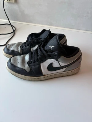 Svarta och vita Air Jordan 1 Low - Säljer ett par Air Jordan 1 Low sneakers i svart och vitt läder med klassisk Nike Swoosh på sidan och Jumpman-logga på plösen. Skorna har svarta snören och en vit sula med svart yttersula. Perfekta för dig som gillar streetwear och ikonisk sneakerstil.
