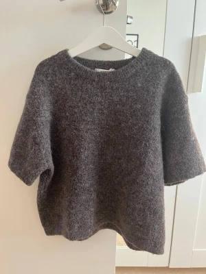 Brun  stickad tröja Gina Tricot - Mysig mörkgrå stickad tröja från Gina Tricot med korta ärmar och rund halsringning. Tröjan har en fluffig, mjuk struktur och är perfekt för lager på lager. Passar dig som gillar en avslappnad och trendig look.