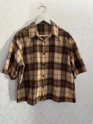 Brun rutig skjorta med tryck - Kortärmad rutig skjorta i bruna och beige toner med stora siffran 07 tryckt på ryggen. Skjortan har klassisk krage, knappar framtill och en loose passform. Perfekt för en avslappnad streetwear-look.