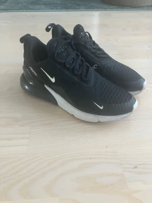 Nike Air Max 270 svarta sneakers - Nike Air Max 270 sneakers i svart med vita detaljer och synlig Air-enhet i hälen. Ovandelen är i mesh för bra ventilation och skon har en chunky, vit mellansula. Klassisk Nike Swoosh på sidan och AIR270-tryck vid hälen. Perfekta för en sportig och trendig look.