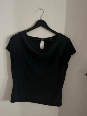 Svart topp med nyckelhålsdetalj - Svart topp från Lindex med korta ärmar och snygg nyckelhålsöppning i nacken. Toppen har en lätt glansig finish och en avslappnad passform som ger en stilren look. Perfekt att styla med jeans eller kjol för en trendig outfit. Snygg och super skön svart topp med veck vid halsen/bröstet. Super skön och skönt tyg. Snygg på. Normal i storlek och bra passform. Det finns små små hål men inget som syns. Storlek S. 