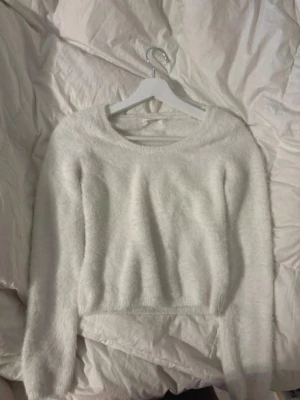 Vit fluffig stickad tröja - Mysig vit stickad tröja med fluffig, mjuk yta och rund halsringning. Tröjan har långa ärmar och ribbade muddar vid ärmslut och nederkant. Perfekt för kalla dagar när du vill vara både varm och snygg.