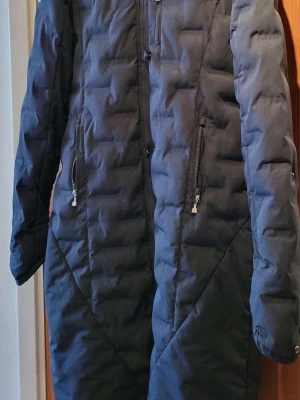 Lång mörkblå dunjacka med huva - Snygg lång mörkblå dunjacka med huva och dragkedja framtill. Jackan har två stora fickor med dragkedja och quiltad design som ger en modern look. Perfekt för kalla vinterdagar och håller dig varm hela dagen.