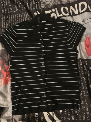 Svart ribbad topp med knappar - Kortärmad svart topp med vita ränder och ribbad struktur. Toppen har knappar framtill och en avslappnad passform. Perfekt till jeans eller kjol för en enkel och stilren look.