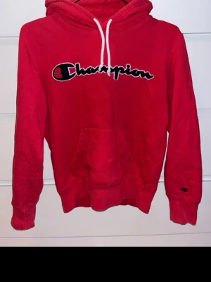 Röd Champion hoodie med logga - Riktigt snygg röd hoodie från Champion med stor broderad logga på bröstet och klassisk känguruficka. Huva med vita snören och ribbade muddar. Perfekt för dig som gillar streetwear och sportig stil. Materialet är mjukt och bekvämt.