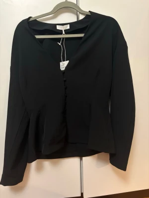 Svart figursydd blus med knappar - Stilren svart blus med figurnära passform och dekorativa klädda knappar framtill. V-ringad hals och långa ärmar ger en elegant look.