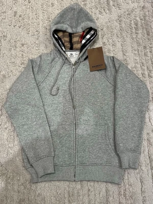 Grå zip hoodie från Burberry - Snygg grå zip hoodie från Burberry med klassiskt rutigt foder i huvan och dragsko. Tröjan har ribbade muddar, fickor framtill och är tillverkad i mjuk bomullsblandning. Perfekt för en chill och stilren look.
