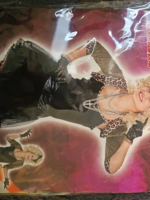 80-tals Madonna maskeradkläder - Snyggt 80-tals Madonna-inspirerat maskeradset med svart klänning, leopardmönstrad bolero, svarta spetsvantar och pärlhalsband. Perfekt för temafest eller cosplay! Klänningen har volangkjol och detaljer i spets och satinliknande material.