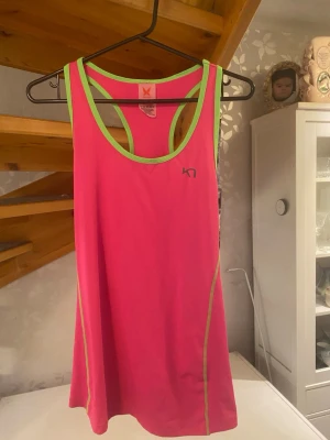 Rosa linnetopp från Kari Traa - Säljer en sportig rosa linnetopp från Kari Traa med gröna detaljer och logga på bröstet. Toppen har brottarrygg och är tillverkad i ett mjukt, stretchigt material som passar perfekt för träning. Snygg kontrast mellan rosa och grönt för en fräsch look.