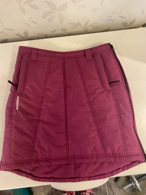Mörkrosa quiltad kjol från Dobsom - Quiltad mörkrosa kjol från Dobsom med dragkedja längs sidan och praktiska fickor framtill. Kjolen har bälteshällor och är något längre bak än fram, perfekt för kalla dagar. Sportig och funktionell design.