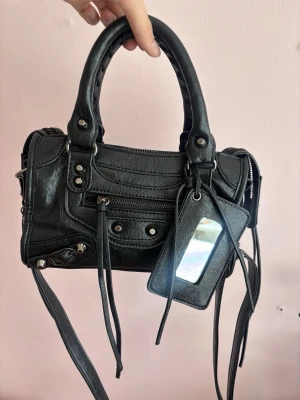 Svart handväska - Söt handväska i läder med silverfärgade nitar och dragkedjor. Väskan har avtagbar axelrem, spegel med inbyggd korthållare samt flera fack på insidan. I fint skick. Pris kan diskuteras 💞