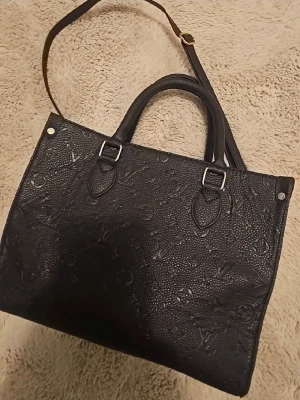 Svart handväska i skinn - Säljer en svart handväska i präglat skinn med klassiskt monogrammönster. Väskan har både handtag och axelrem, silverfärgade metalldetaljer och en stilren, fyrkantig form. Perfekt för dig som vill ha en lyxig och tidlös accessoar.