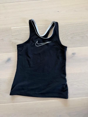 Svart Nike träningslinne - Säljer ett svart träningslinne från Nike med material som håller dig torr under träningen. Linnet har en racerback-design och vita detaljer längs kanterna samt Nike-logga på bröstet. Perfekt för gym, löpning eller sport🔥🔥